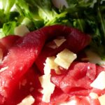 Bresaola appoggiata su un tagliere di legno, con fette sottili e decorazioni di rucola e pomodorini.