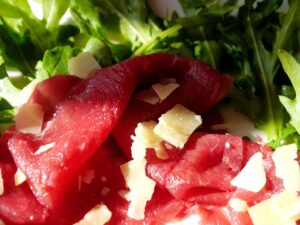 Bresaola appoggiata su un tagliere di legno, con fette sottili e decorazioni di rucola e pomodorini.